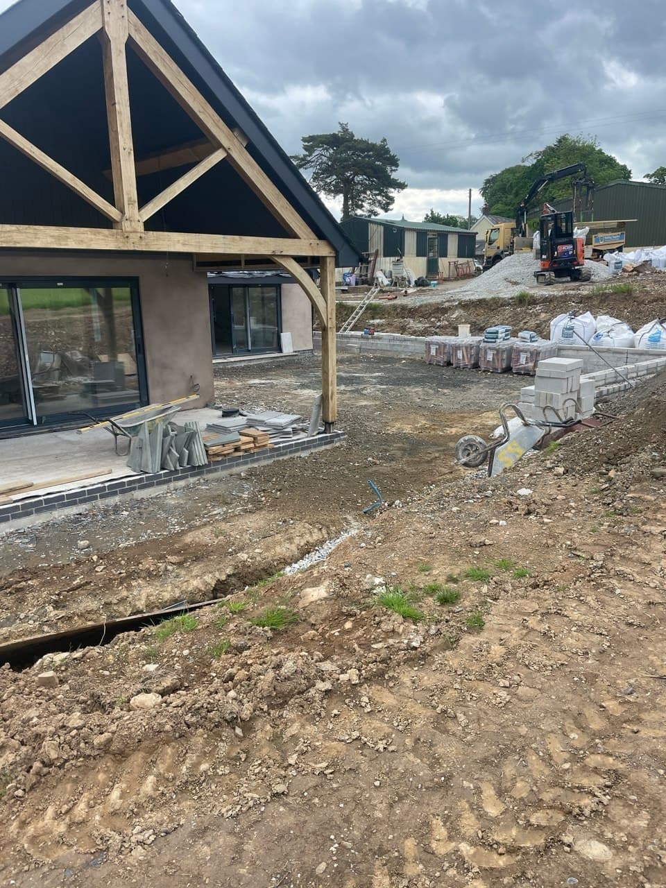 Oak-framed carport gable end with mini excavator and groundworks in progress at Thornbury Devon new build site